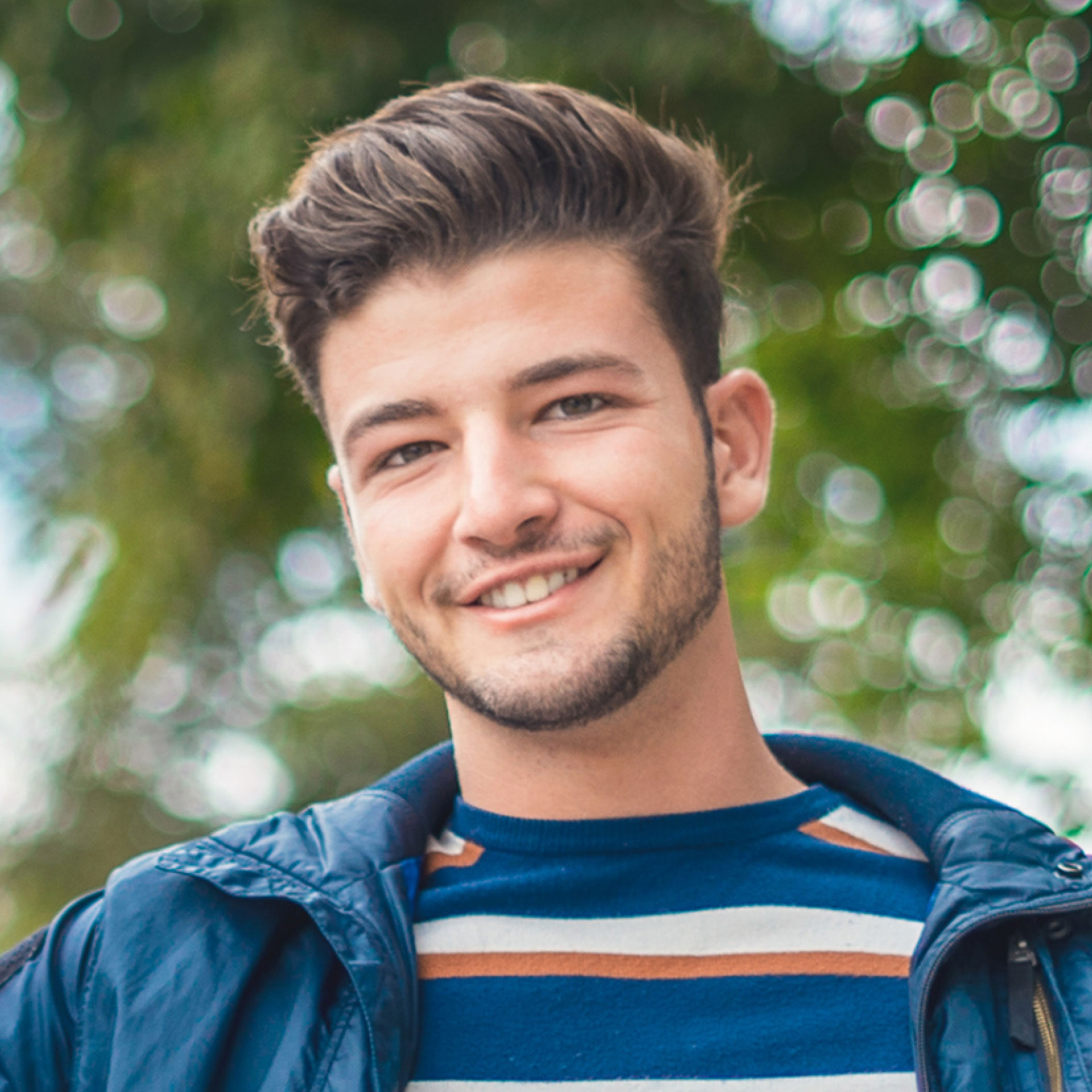 young man wearing striped shirt and denim jacket sitting outside smiling at camera