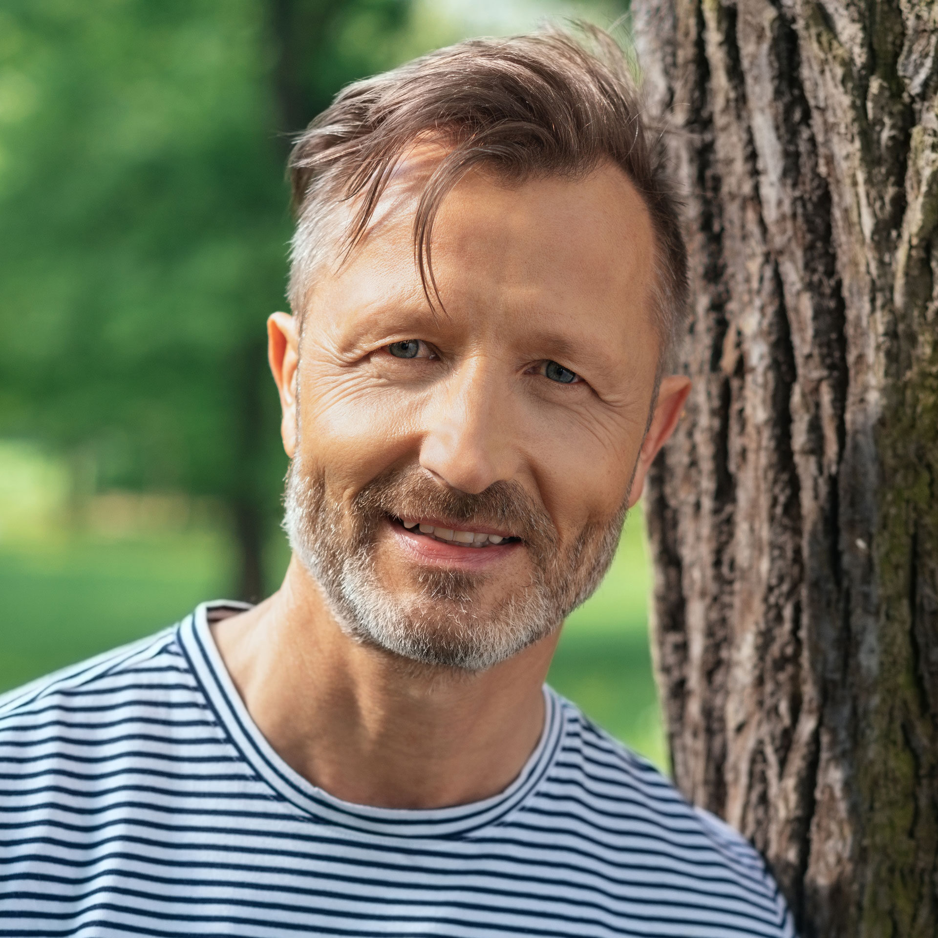middle-aged man wearing striped shirt leaning against tree smiling at camera